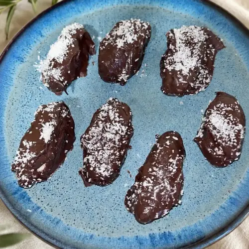 Sieben hausgemachte, vegane Bounty-Riegel mit Schokoladenüberzug und Kokosstreuseln auf einem blauen Keramikteller.